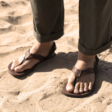 Person wearing brown leather flip-flops standing on sandy beach, with olive green rolled-cuff pants.