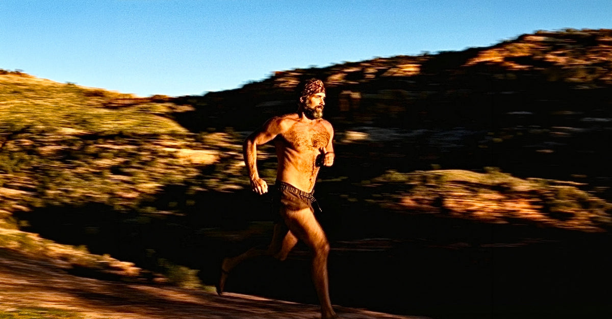 A bearded man wearing a bandana, shirtless, dark running shorts, and running sandals sprints along a dirt trail through a sun-drenched desert landscape. He is captured mid-stride with motion blur in the background conveying speed. Rocky hills with sparse desert vegetation are lit by warm golden light, with a clear blue sky above. The dramatic panning photography technique keeps the subject in focus while the landscape streaks behind him.